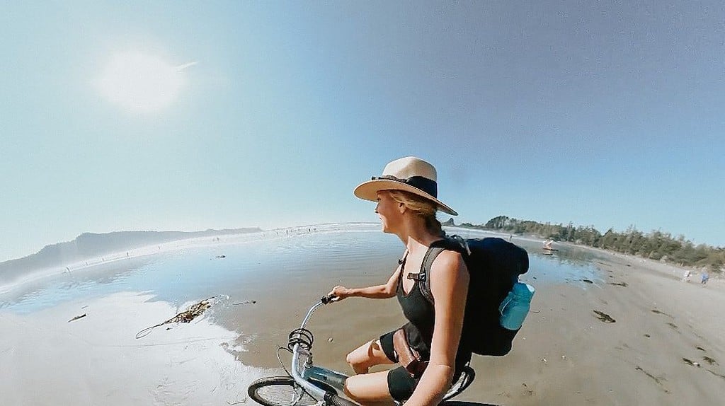 A white woman wearing a hat is riding a cruiser bike along the beach in Tofino BC